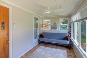 Quarto com porta de madeira, sofá azul com almofada marrom, tapete branco, ventilador de teto e janela com persianas.