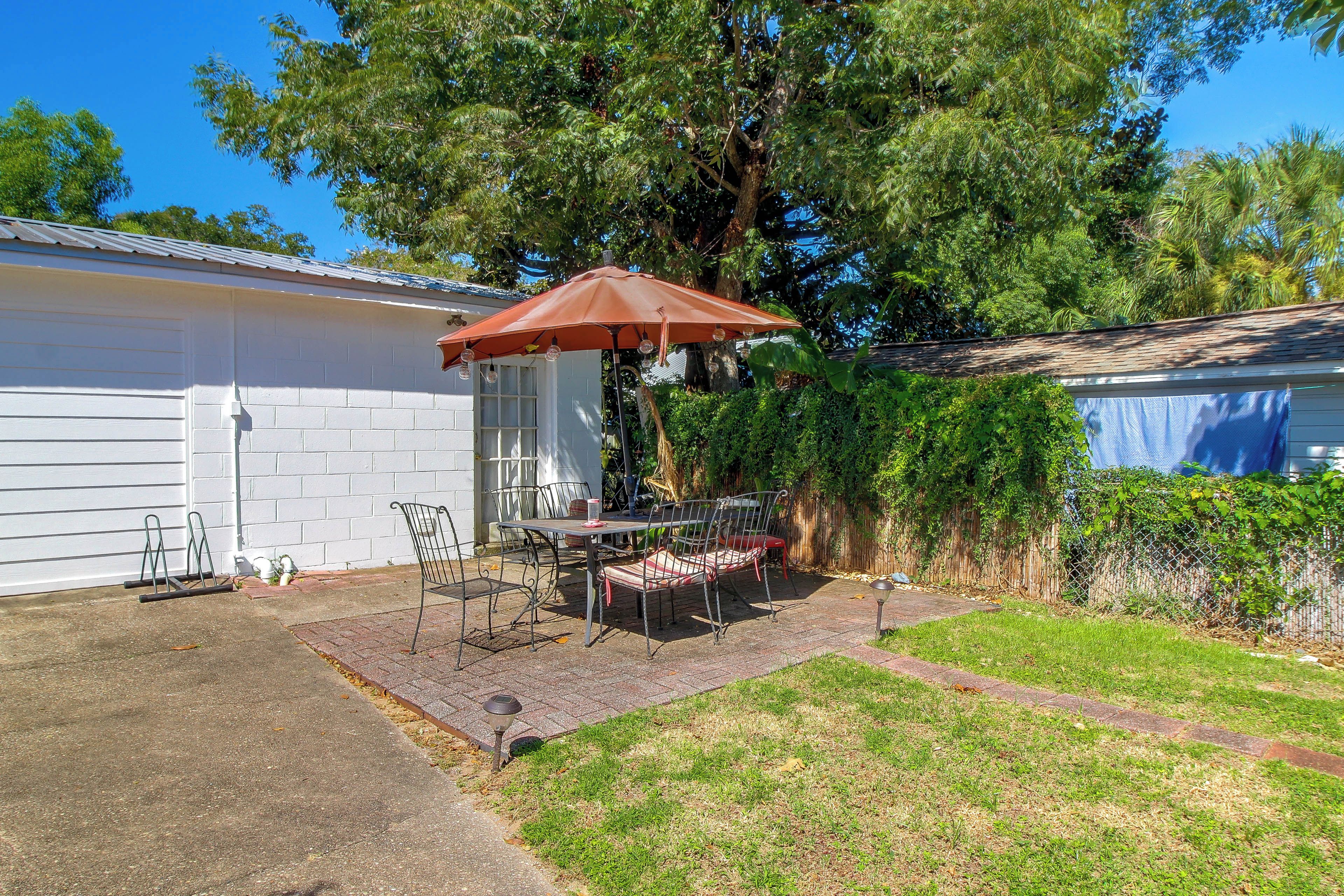 A patio with a table and chairs, a red umbrella, and a white building in the background.