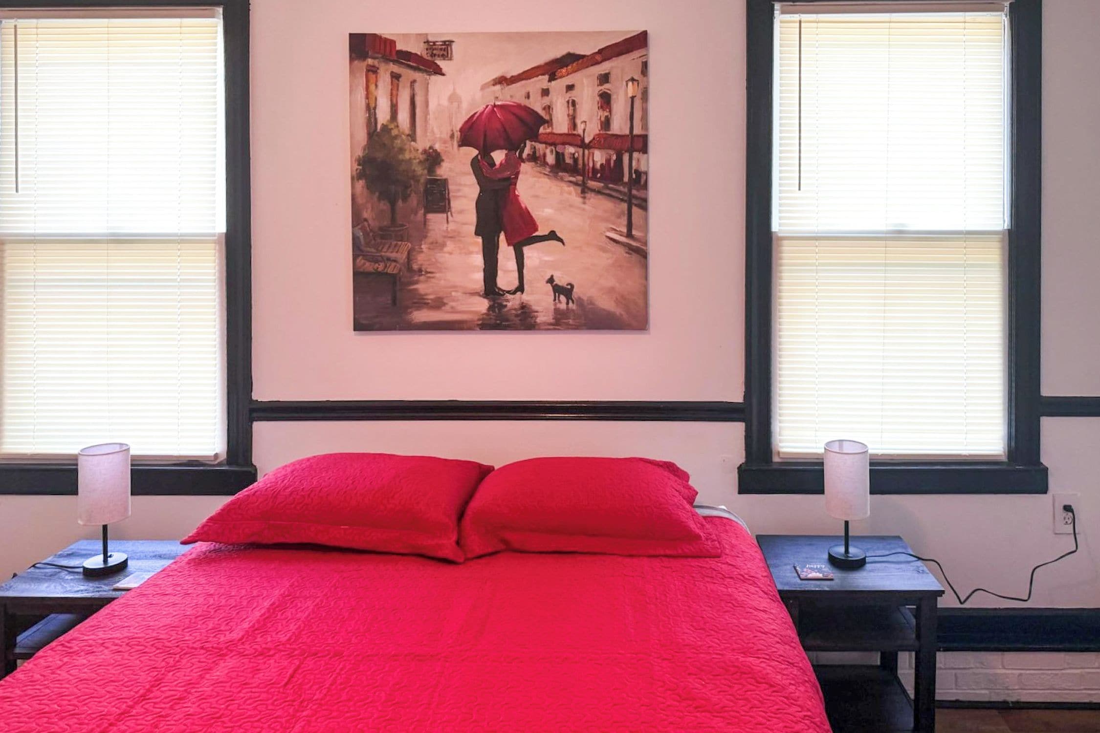 A bedroom with a bed, two bedside lamps, a painting on the wall, and windows with blinds.