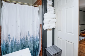 Casa de banho com cortina de chuveiro estampada com uma floresta, porta-toalhas com toalhas dobradas e chão de madeira.
