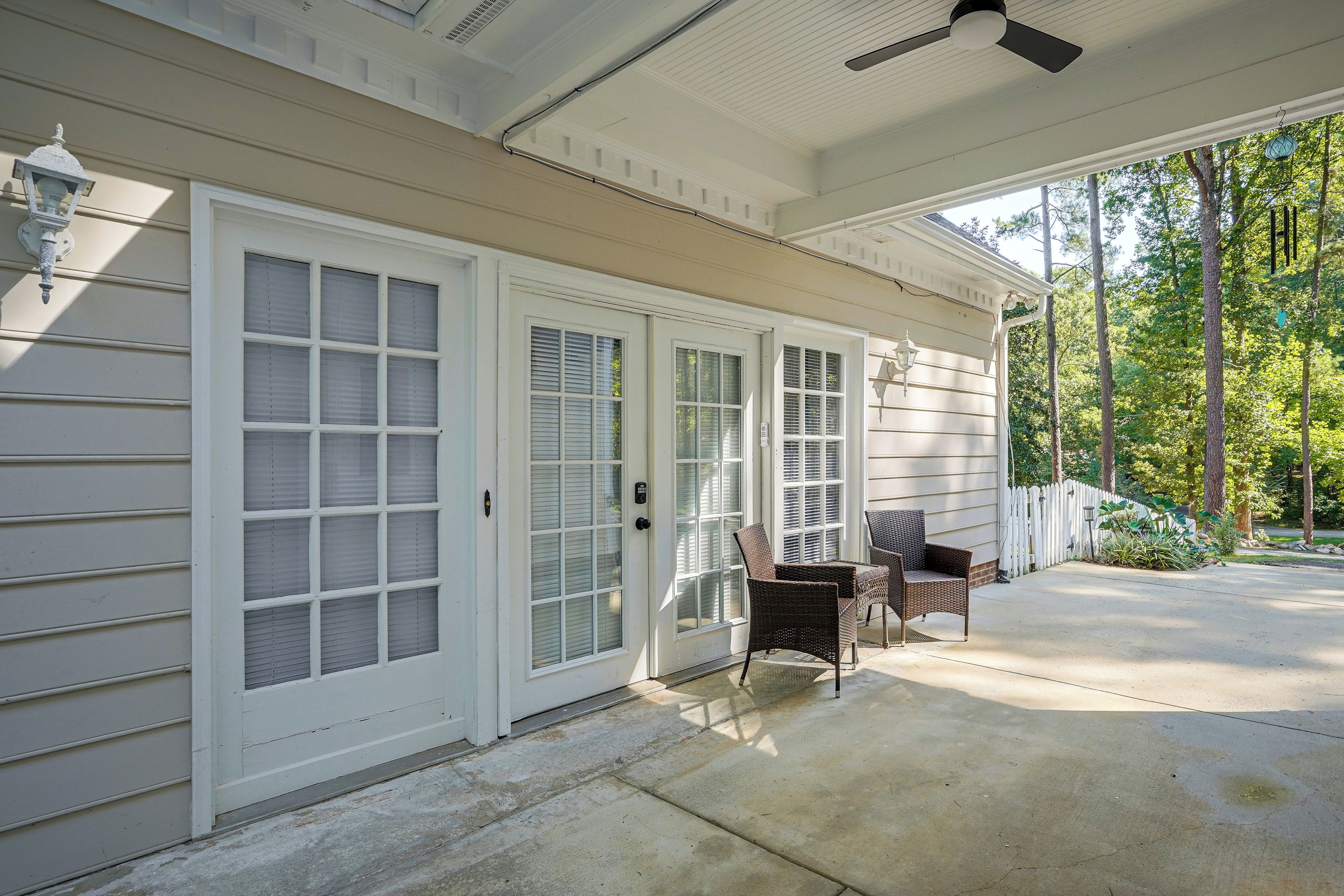 A covered porch with two wicker chairs, a white door with glass panels, and a white fence.