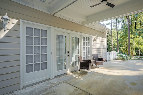 A covered porch with two wicker chairs, a white door with glass panels, and a white fence.