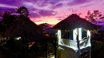 A thatched-roof structure with a staircase leading to a covered area, under a twilight sky.