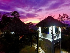 A thatched-roof structure with a staircase leading to a covered area, under a twilight sky.