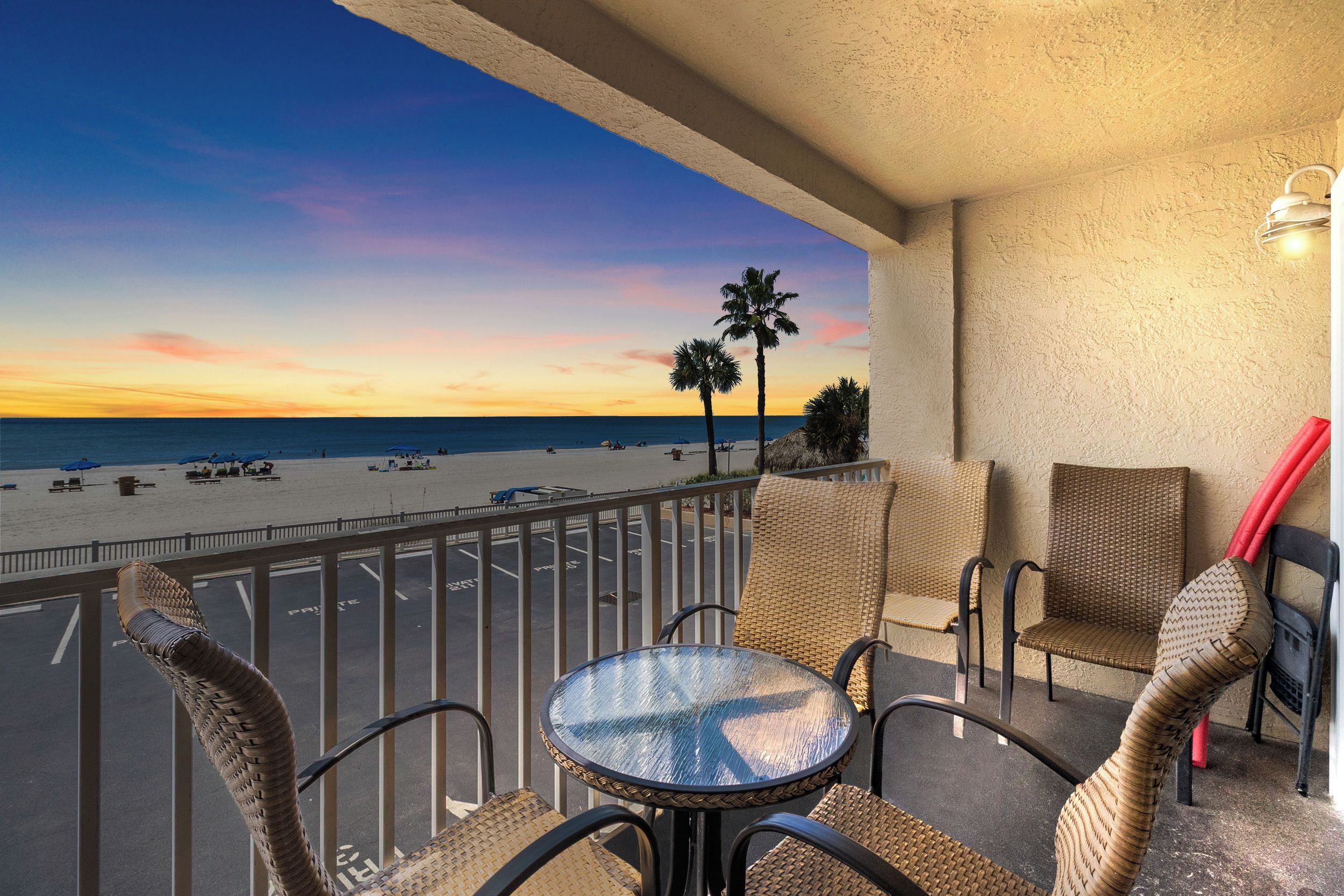 Un balcon agrémenté de meubles en osier, d’une table en verre et offrant une vue sur la plage et l’océan au coucher du soleil.