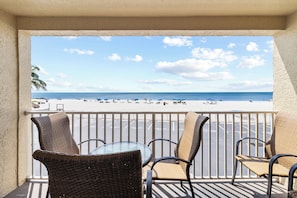 Ein Balkon mit Korbstühlen und einem Tisch, der Blick auf einen Strand mit Personen und Sonnenschirmen bietet.