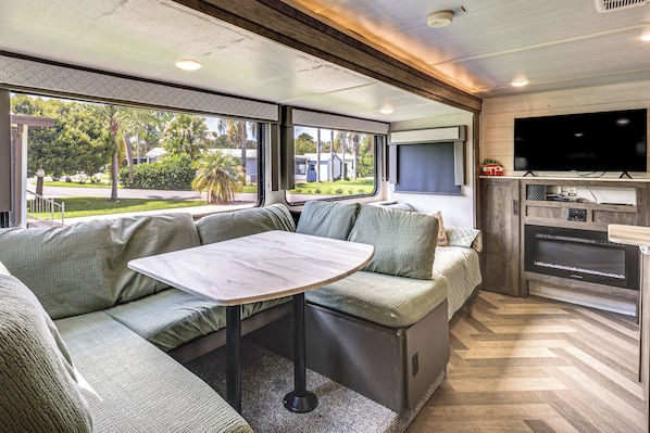 A modern RV interior with a dining area, seating, a television, and a kitchen area.