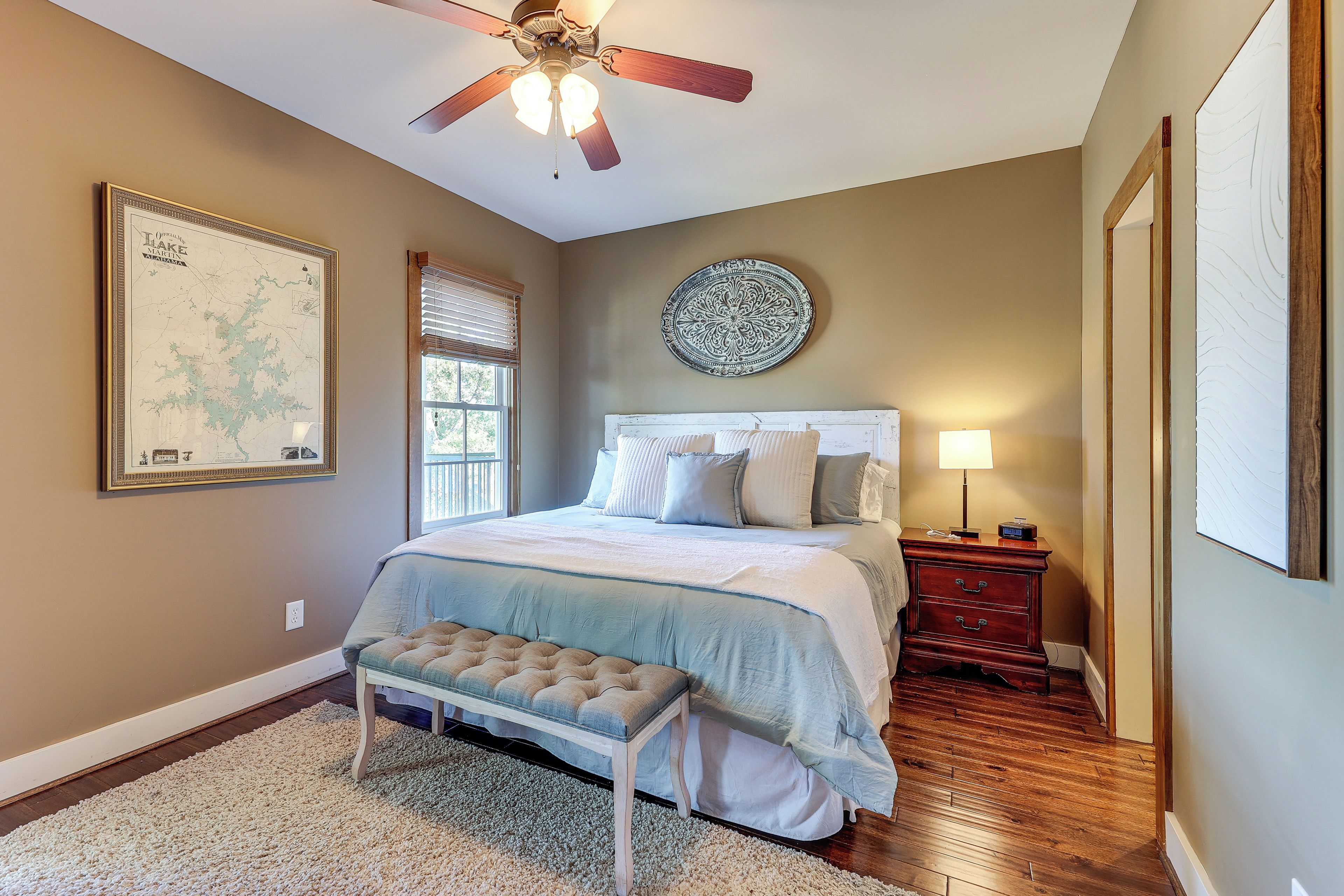 A bedroom with a bed, a ceiling fan, a nightstand, a lamp, a map on the wall, and a window with blinds.