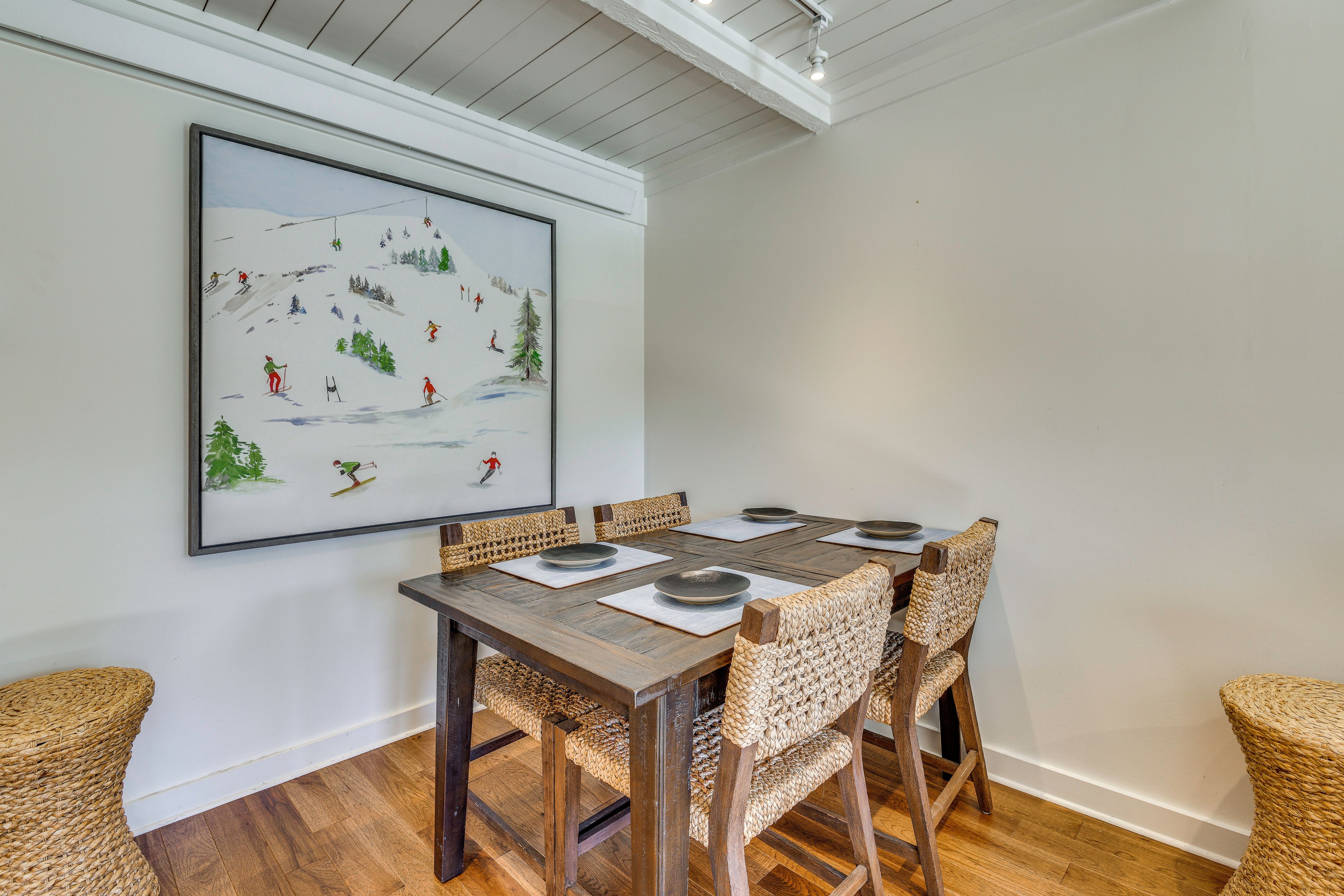 A dining area with a wooden table, woven chairs, and a framed painting of a ski scene.