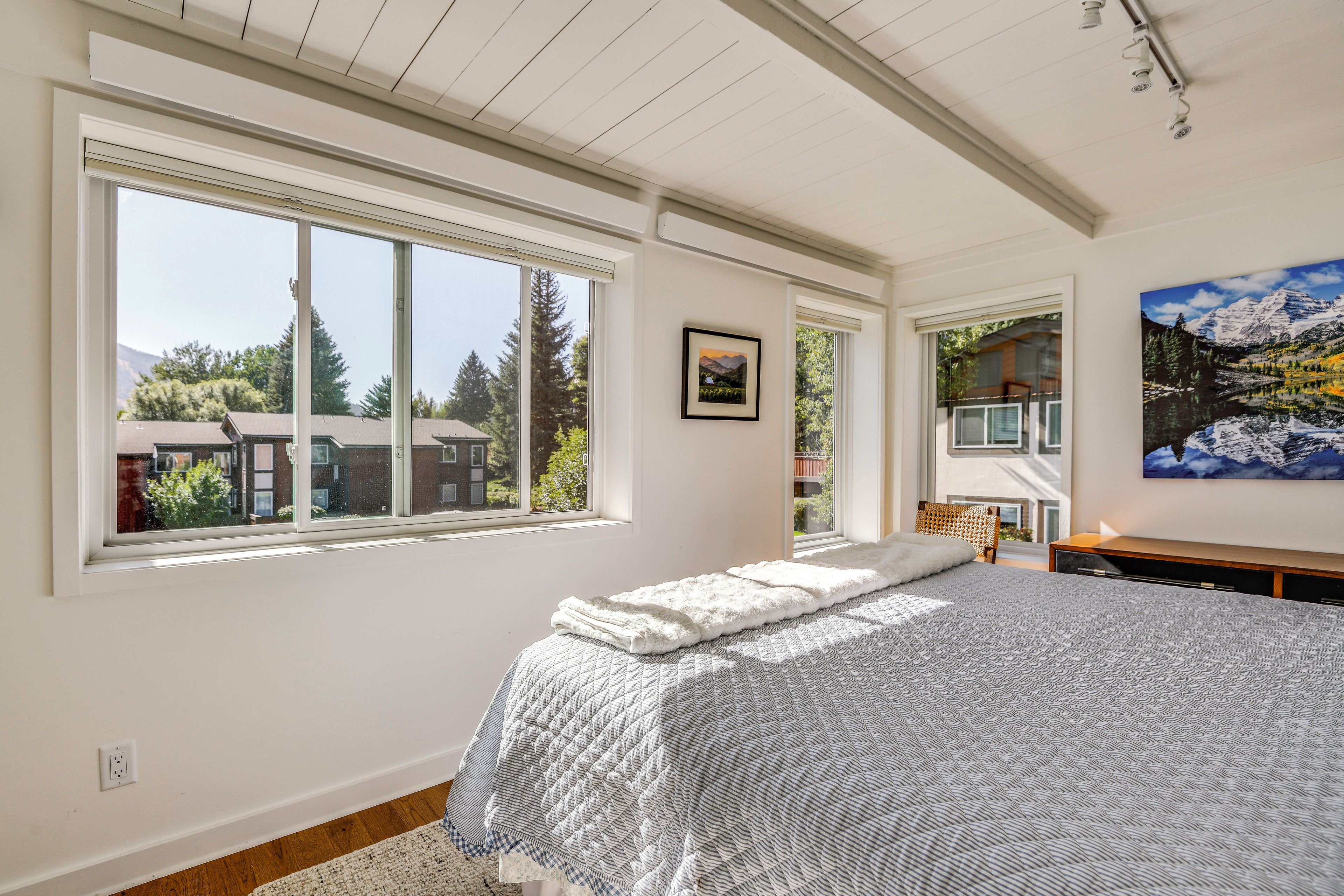A bedroom with a large window, a bed with a white cover, a painting, and a view of a mountain.