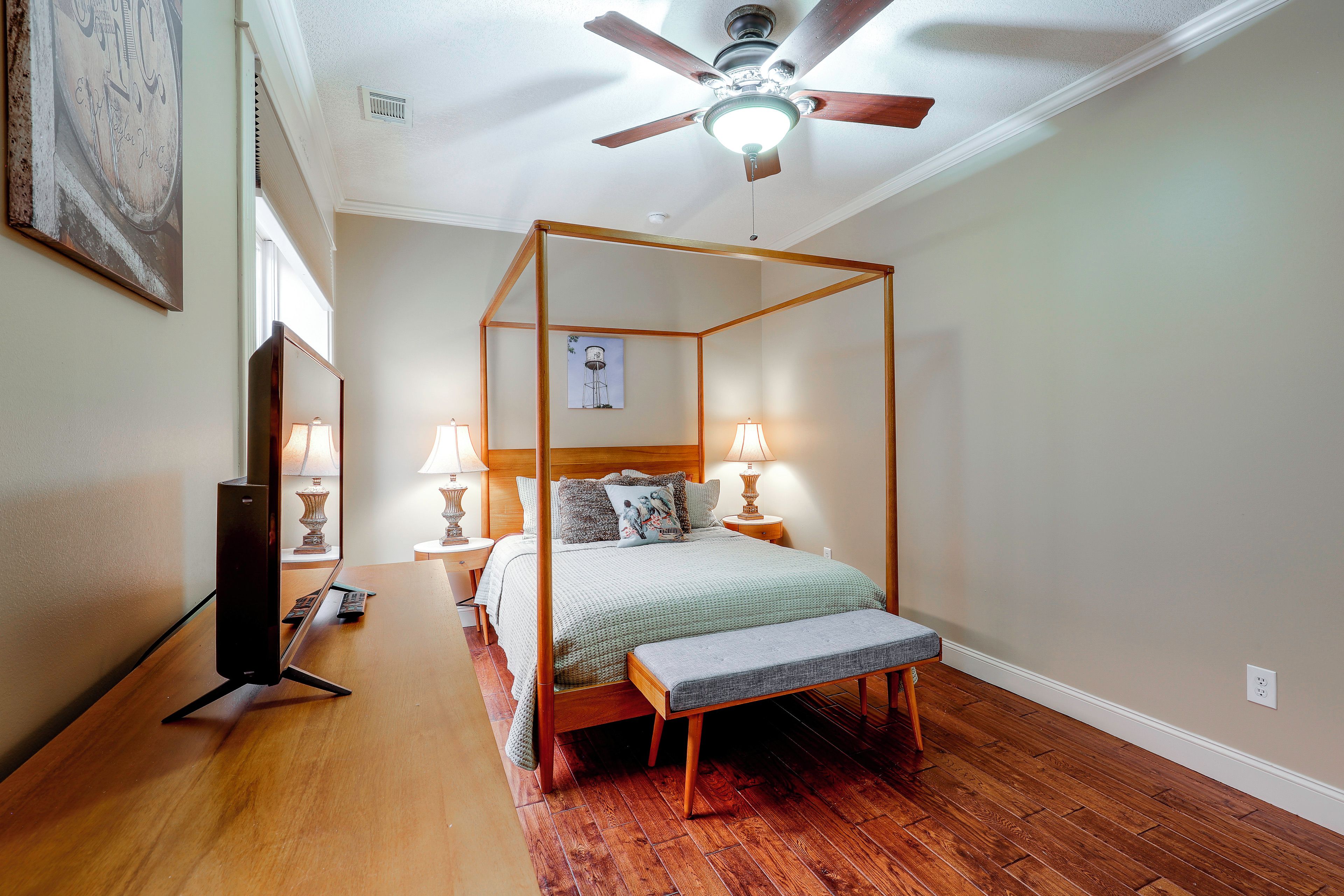 A four-poster bed with a wooden frame, two bedside tables with lamps, a ceiling fan, and hardwood flooring.