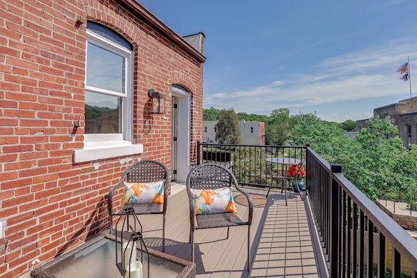 A brick building with a balcony featuring two wicker chairs with cushions, a small table, and a railing.