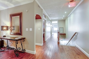 A hallway with wooden flooring, a mirror, a table with a lamp, and an exit sign.