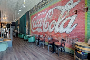 Un interior de restaurante con un gran mural de Coca-Cola en la pared, suelo de madera y asientos tapizados en verde.