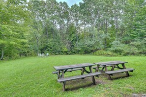 Un área de picnic con bancos y mesas de madera, rodeada por un campo cubierto de césped y árboles frondosos.