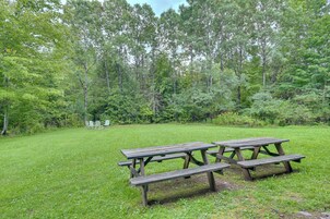 Un área de picnic con bancos y mesas de madera, rodeada por un campo cubierto de césped y árboles frondosos.