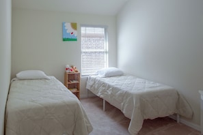 A room with two single beds, a window with blinds, a small shelf with books and toys, and a wall decoration. - Lewisville Lake Escape, 4 Mi to Boat Ramp (Little Elm)