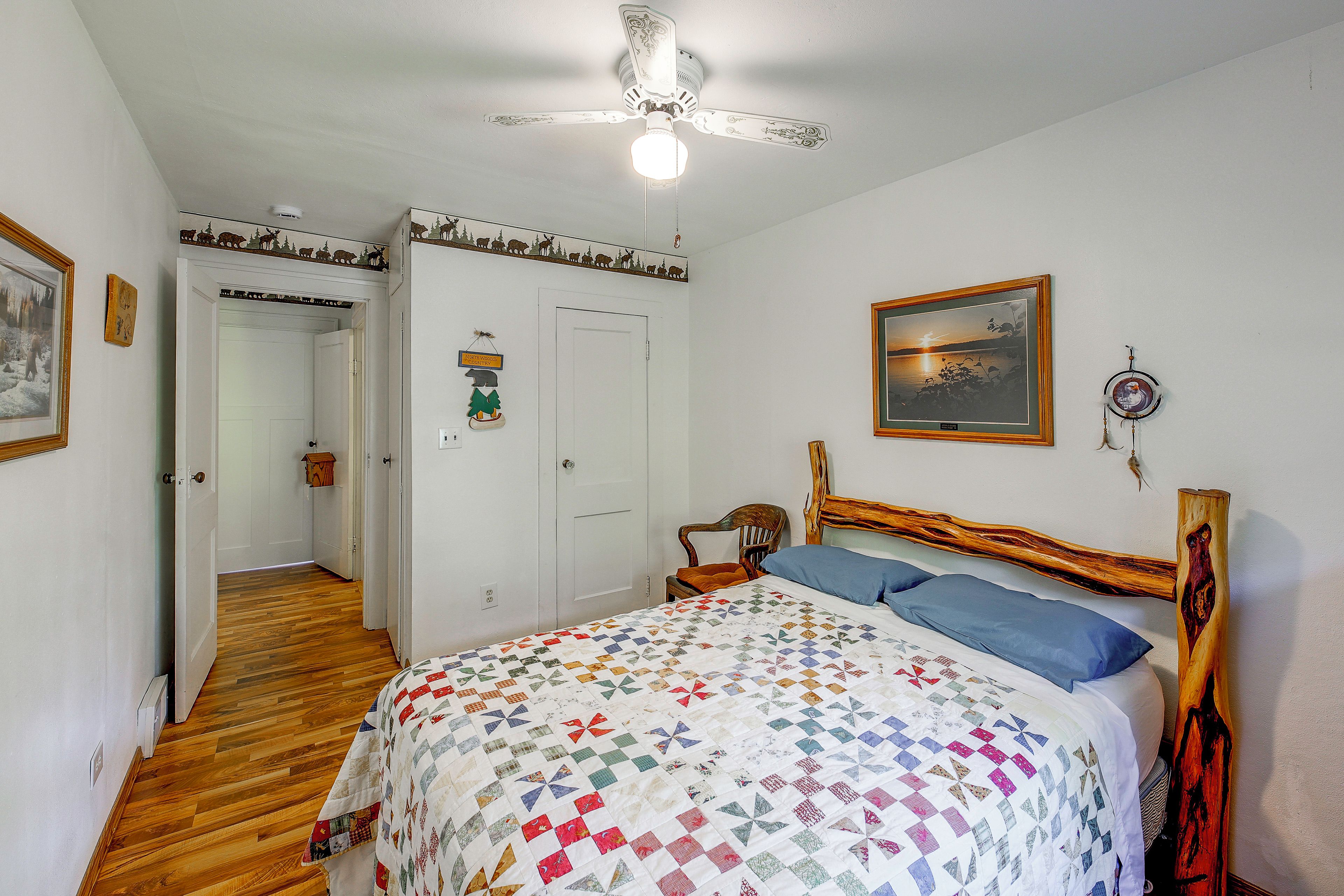 A bedroom with a wooden floor, a bed with a colorful quilt, a ceiling fan, and a framed picture on the wall.