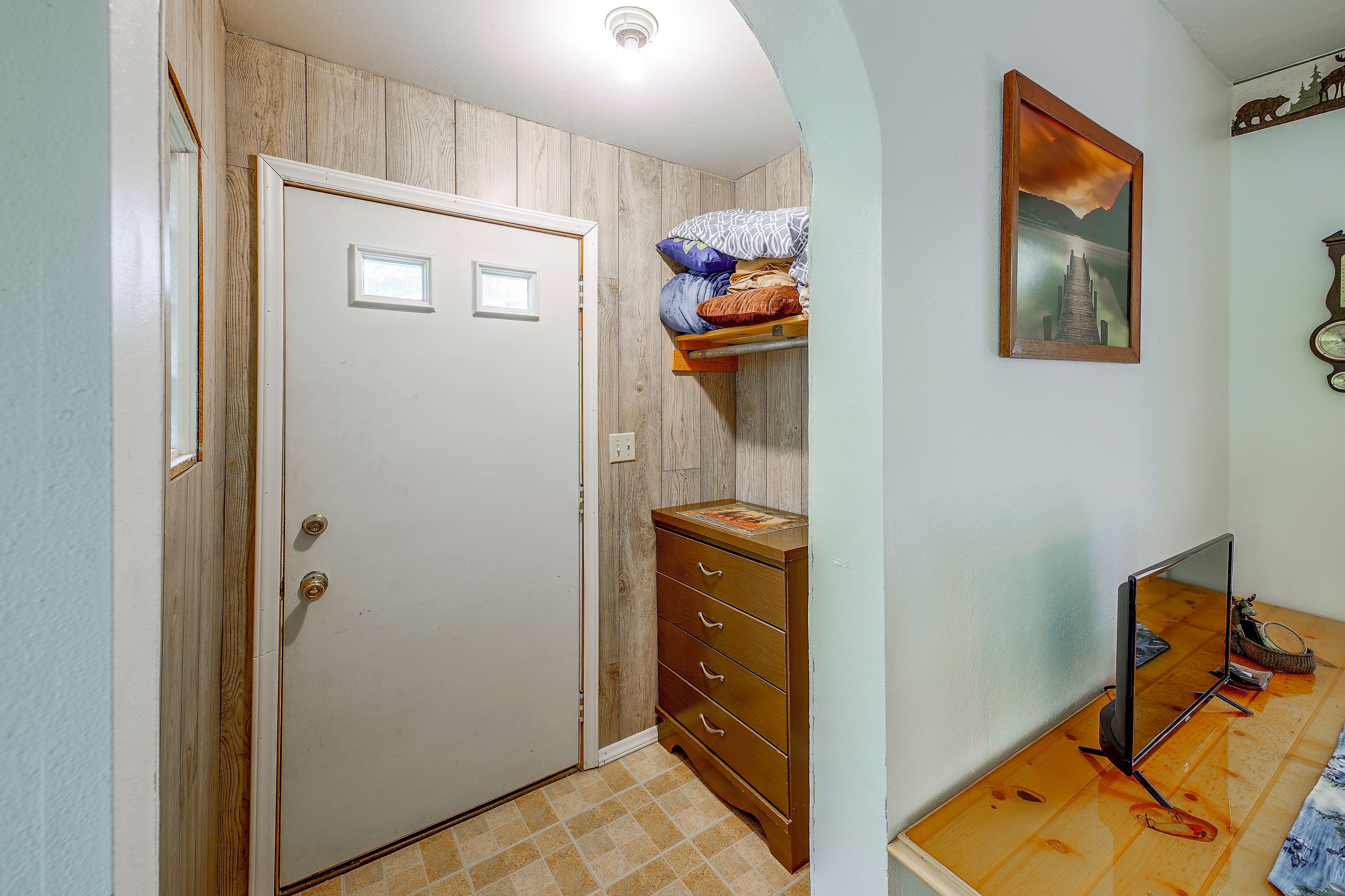 Un pasillo con puerta de madera, cómoda con cajones, estante con almohadas, un cuadro enmarcado en la pared y un espejo en el suelo.