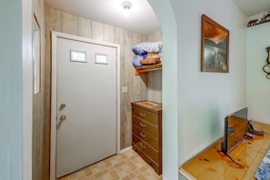 A hallway with a wooden door, a dresser with drawers, a shelf with pillows, a framed picture on the wall, and a mirror on the floor.