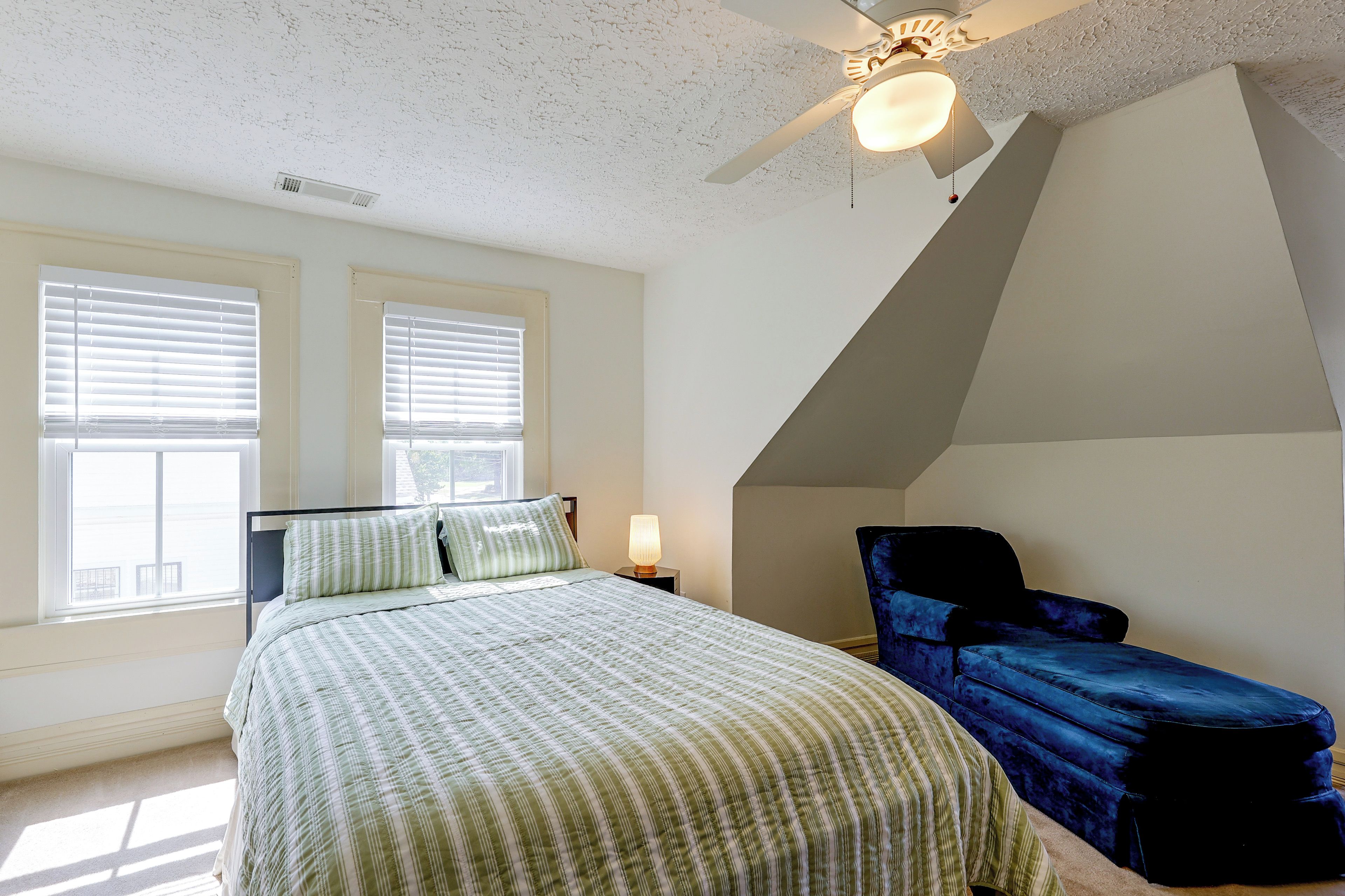 A bedroom with a bed, two windows, a ceiling fan, and a blue chair.