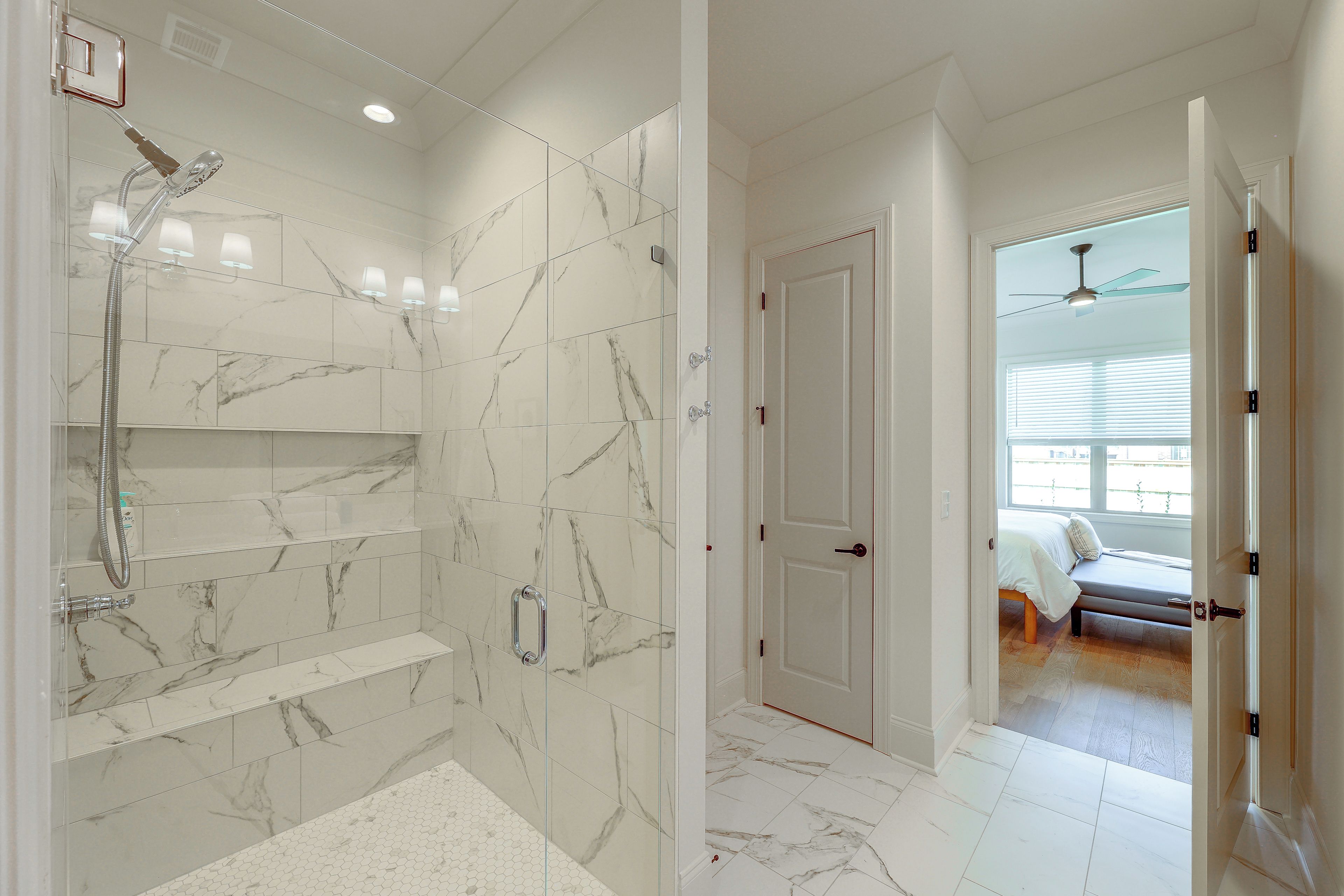 A modern bathroom with a glass-enclosed shower, marble tiles, and a visible door leading to a bedroom with a bed and window.