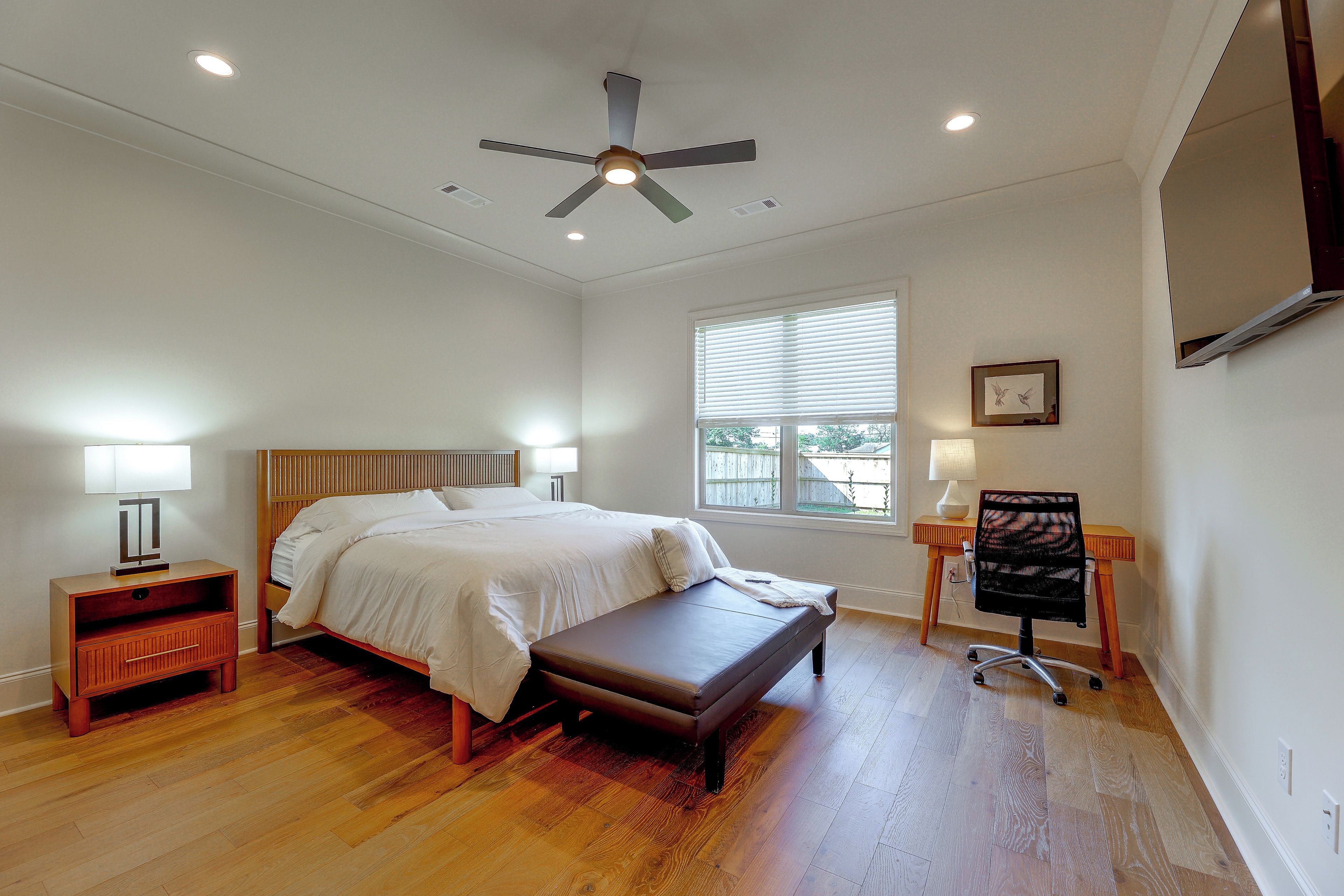 A bedroom with a bed, bedside tables, a bench, a desk with a chair, a ceiling fan, and a window with blinds.