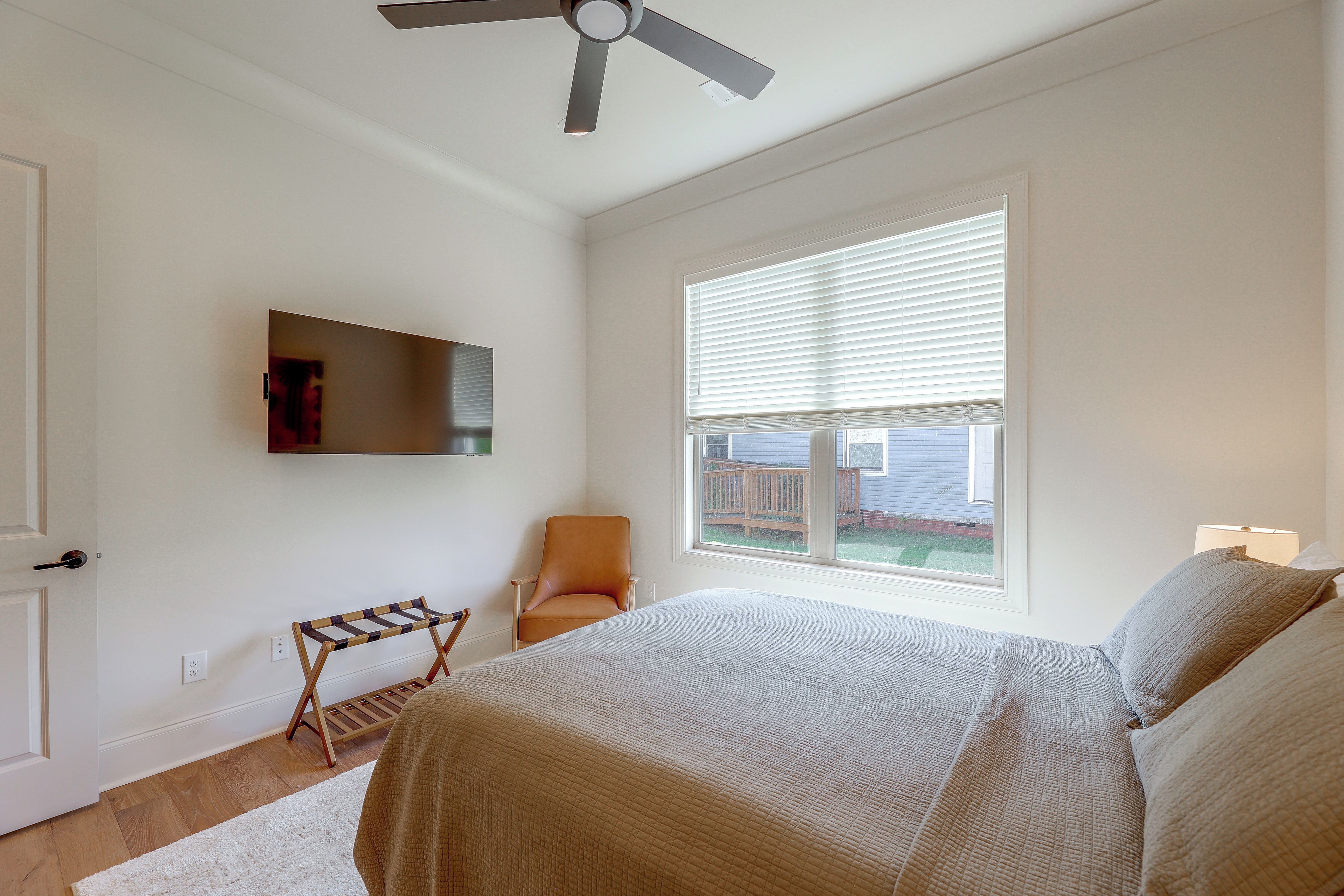A bedroom with a bed, a chair, a TV, a window with blinds, and a ceiling fan.