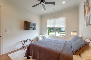Un dormitorio con cama, ventilador de techo, televisor montado en la pared, ventana con persianas y una pequeña mesa con silla.