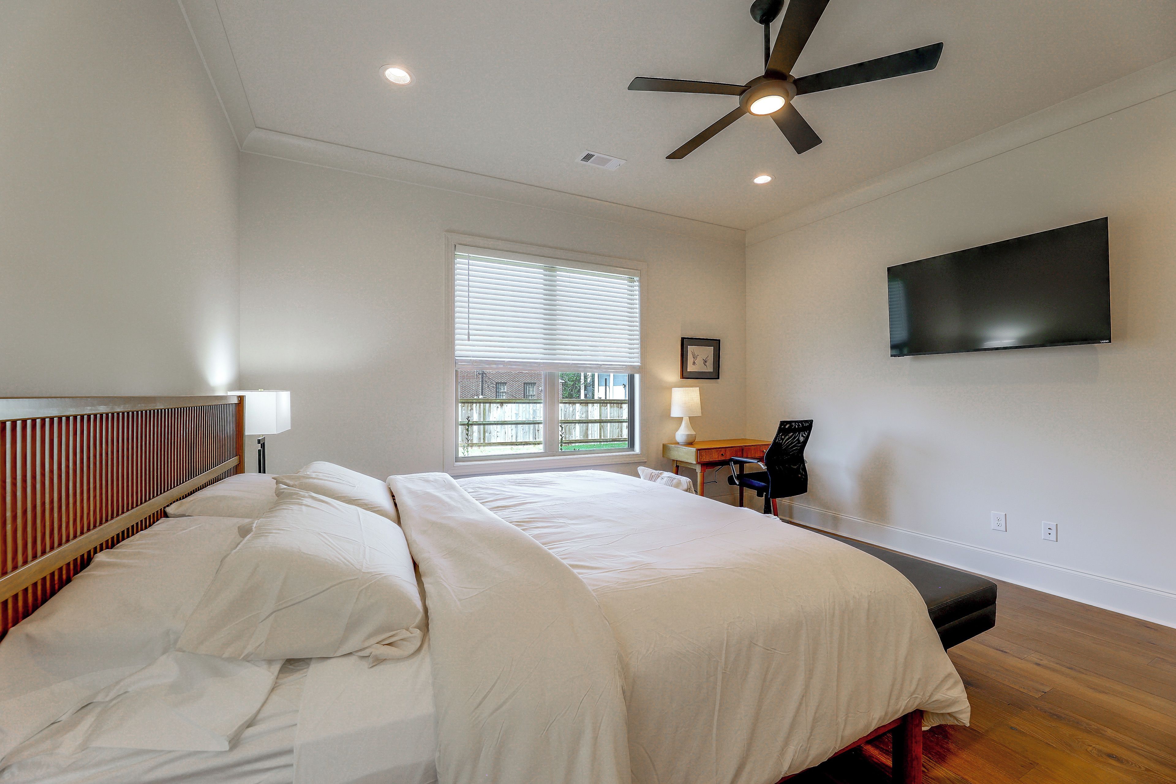 A bedroom with a bed, a ceiling fan, a window with blinds, a bedside table with a lamp, and a flat-screen TV mounted on the wall.