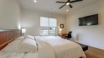 A bedroom with a bed, a ceiling fan, a window with blinds, a bedside table with a lamp, and a flat-screen TV mounted on the wall.
