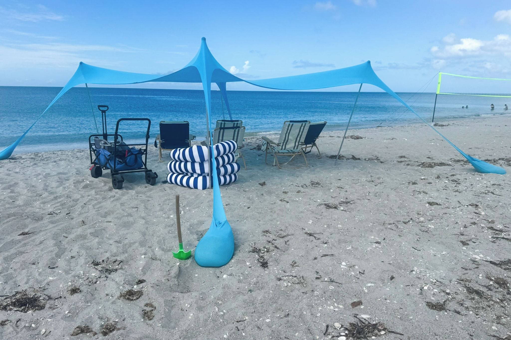 Una playa con una estructura tipo sombrilla azul, sillas de playa y un carrito con bolsas.