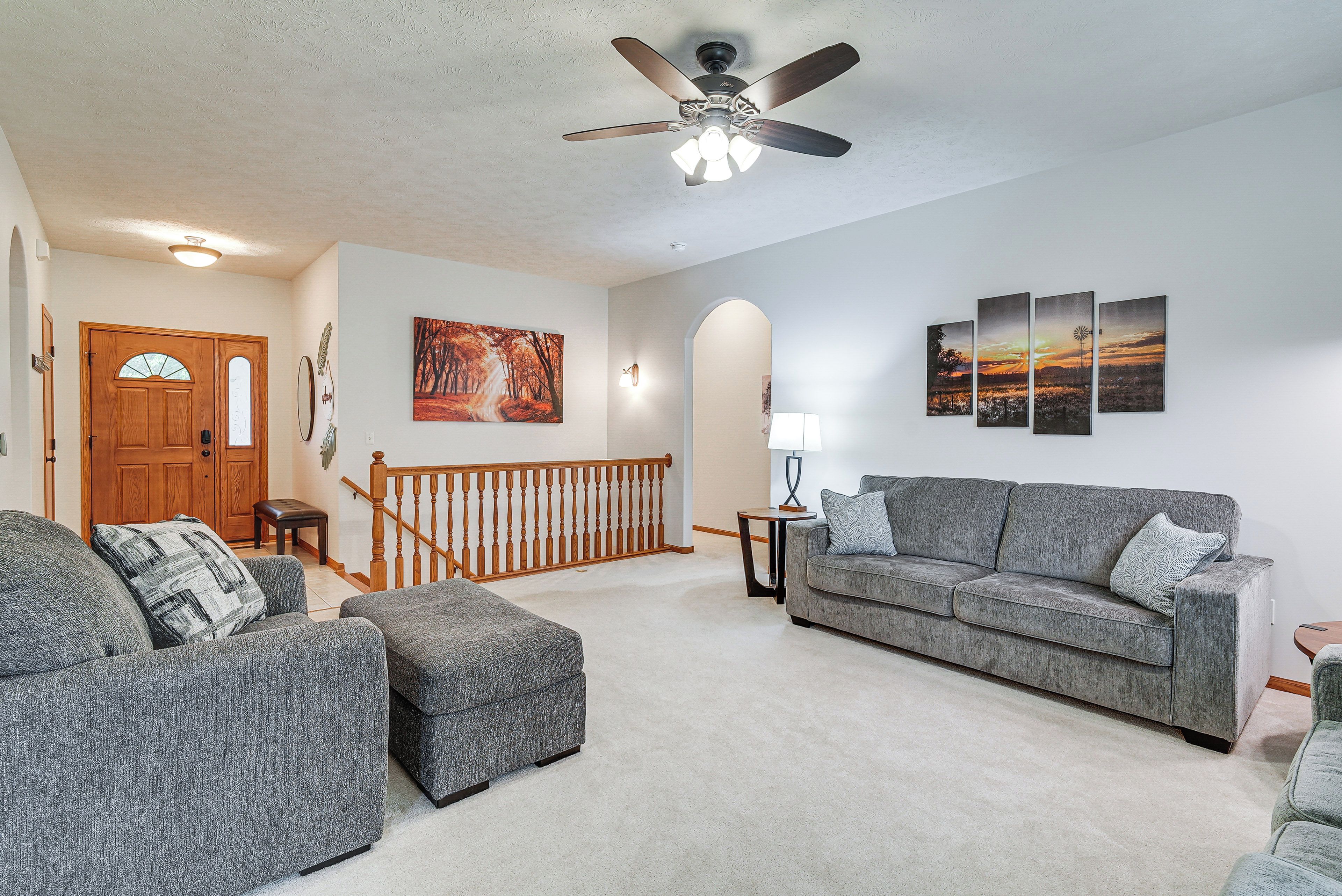 A living room with a grey sofa, two grey armchairs, a wooden staircase, and a ceiling fan.