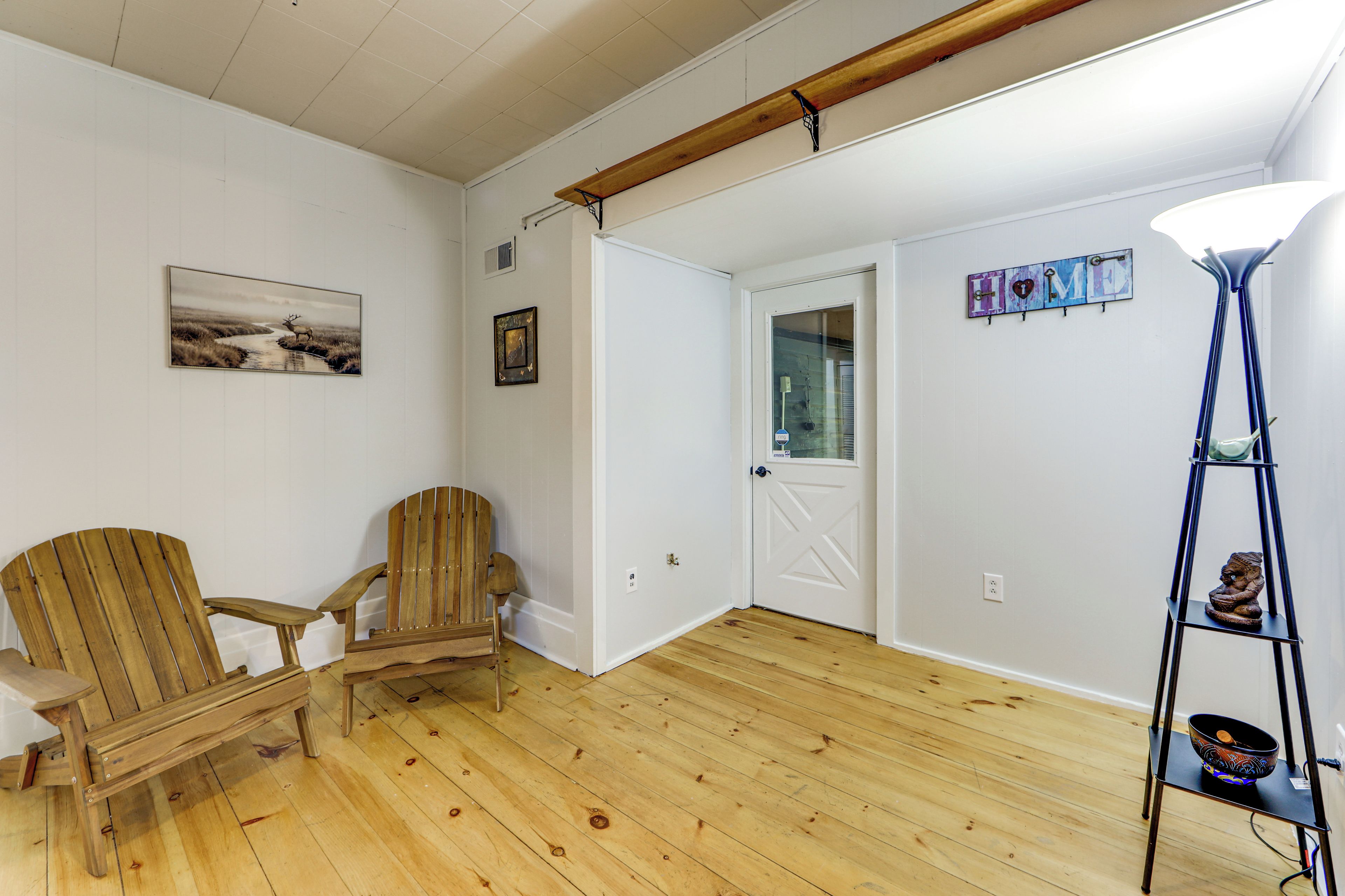 A room with wooden flooring, a wooden chair, a small table, a lamp, and a wall-mounted shelf with a decorative item.