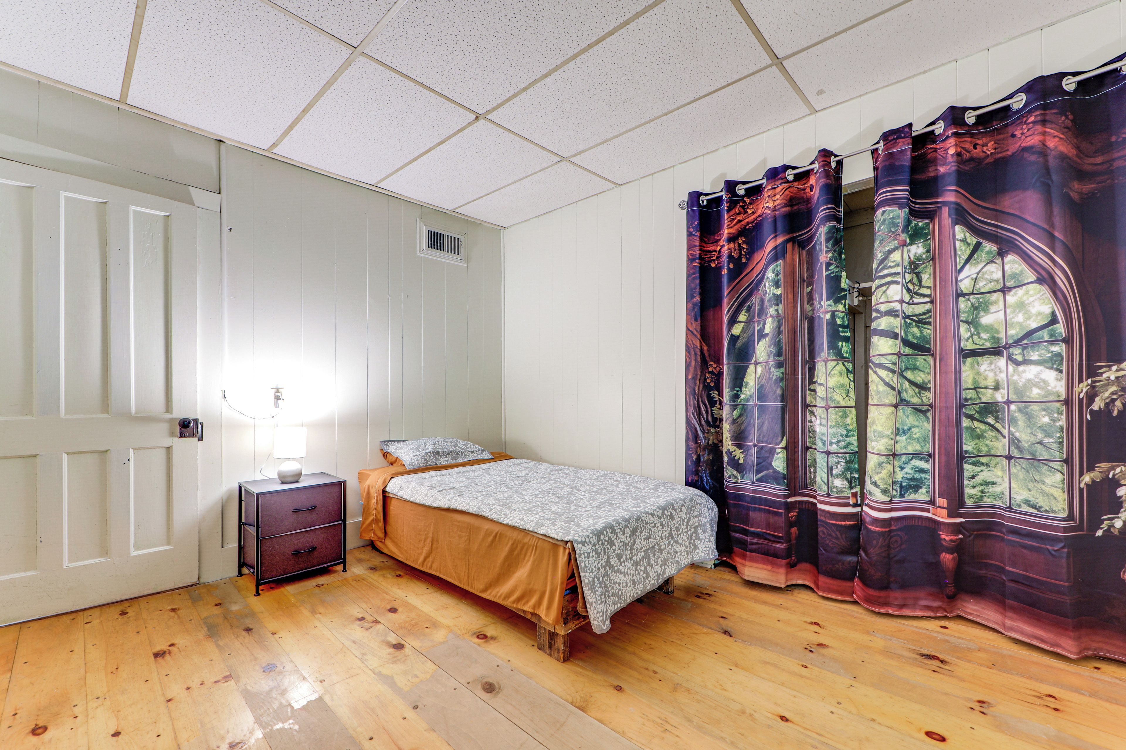 A bedroom with a single bed, a nightstand, a lamp, a window with curtains, and a door.