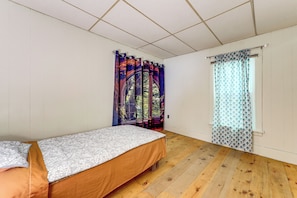A bedroom with a single bed, wooden flooring, a window with patterned curtains, and a door with floral curtains.