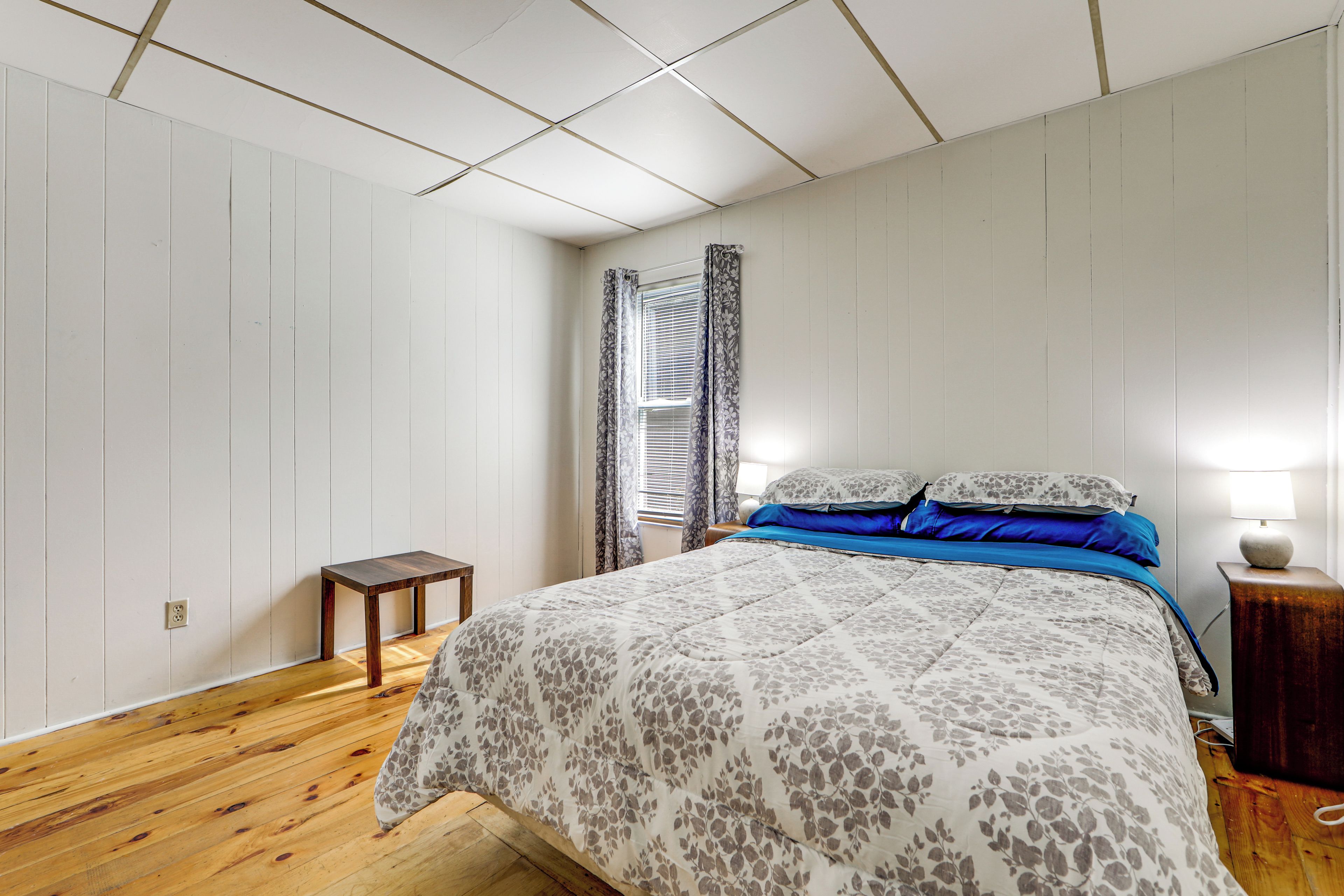 Un dormitorio con piso de madera, una cama con ropa de cama floral, una mesita de noche con lámpara y una ventana con cortinas.
