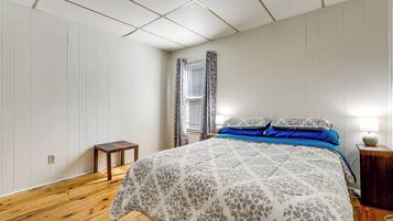 Un dormitorio con piso de madera, una cama con ropa de cama floral, una mesita de noche con lámpara y una ventana con cortinas.