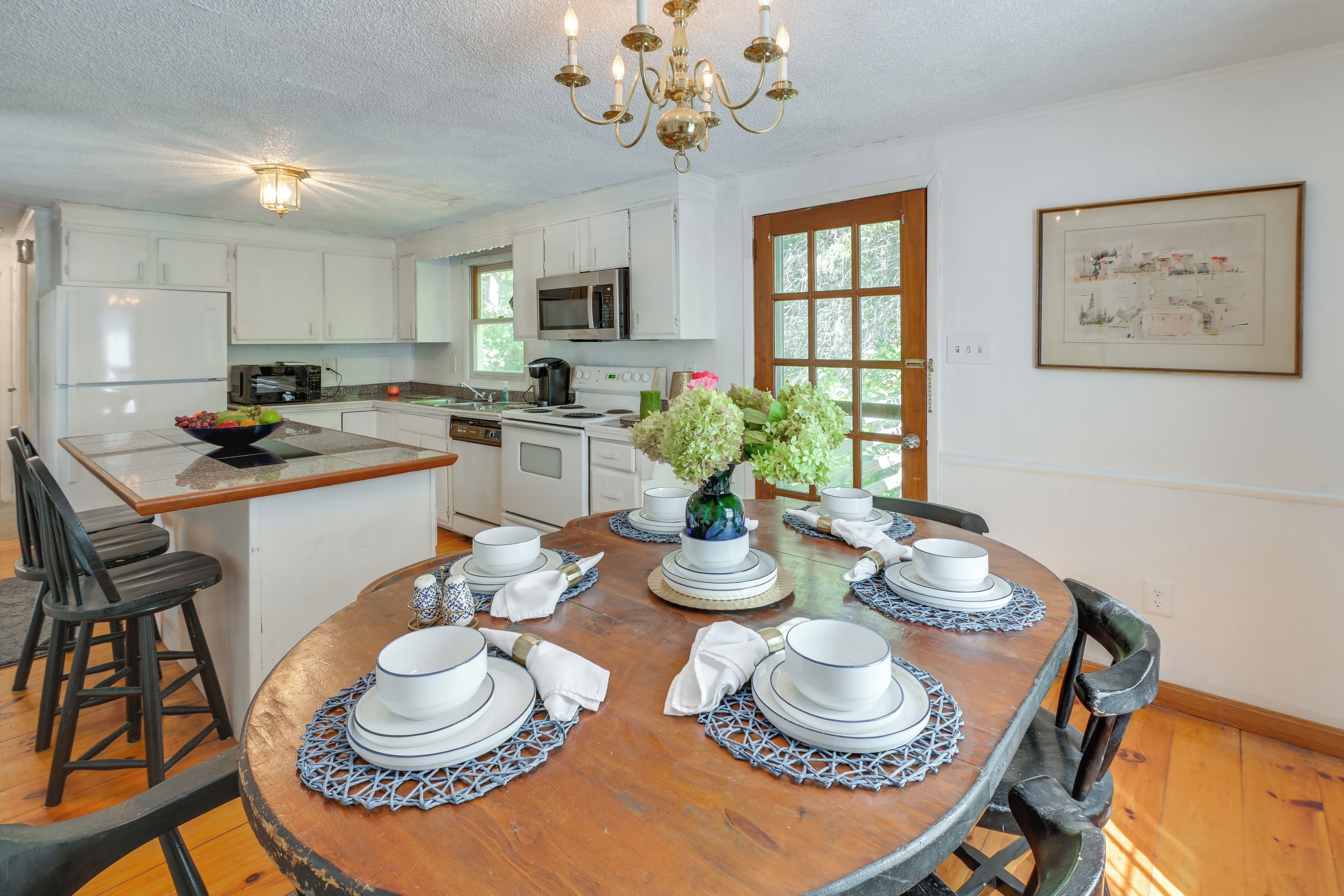 A modern kitchen with white cabinets, a microwave, and a sink. There's a dining table set with plates, cups, and a vase of flowers.