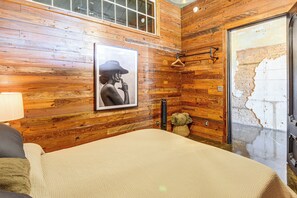 A room with wooden panel walls, a bed with a beige cover, a framed picture on the wall, a towel rack, and a teddy bear.