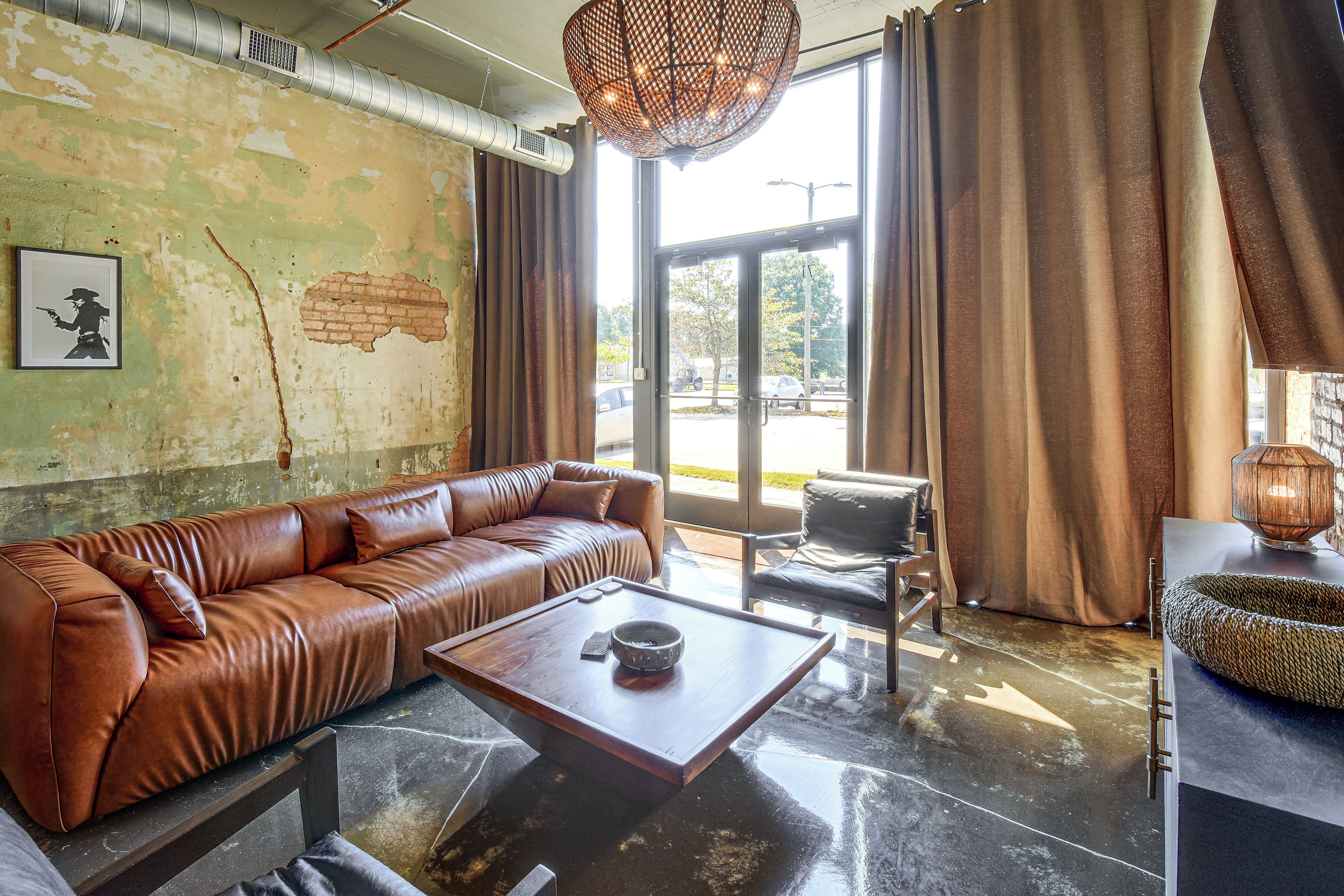 A room with a brown leather sofa, a coffee table, a wicker basket, and a large window with curtains.