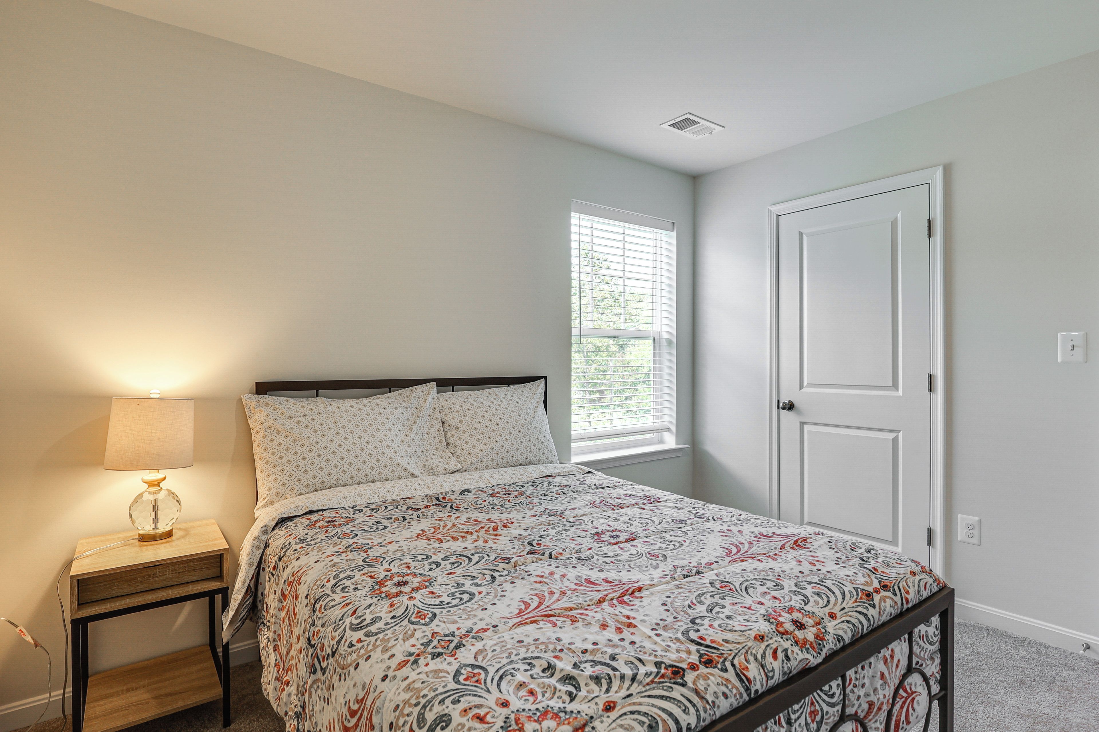 A bedroom with a bed, a nightstand, a lamp, a window with blinds, and a door.