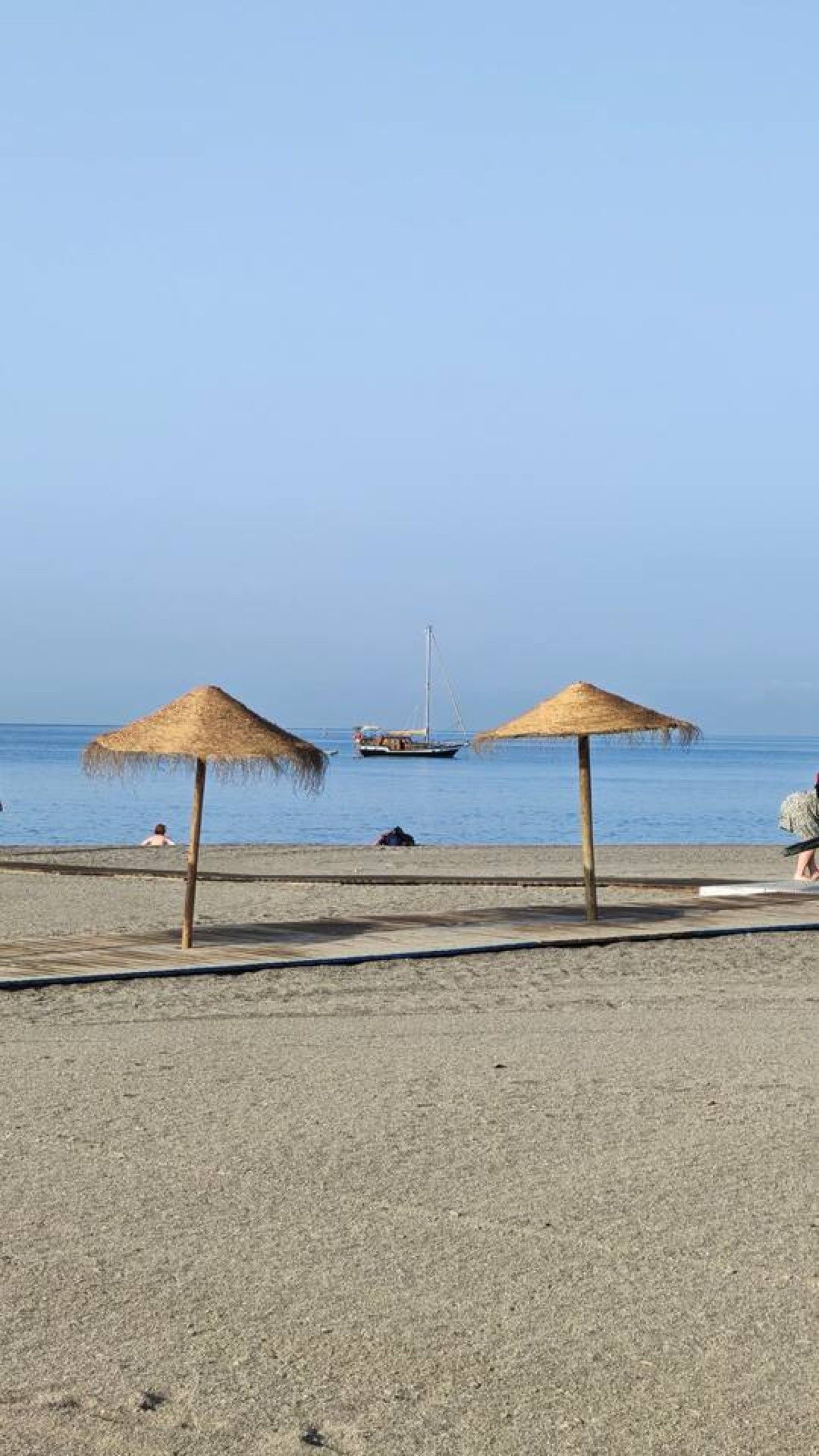 Une plage avec deux parasols en chaume, un rivage sablonneux et un voilier à l’horizon.