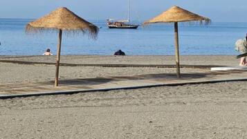 Une plage avec deux parasols en chaume, un rivage sablonneux et un voilier à l’horizon.