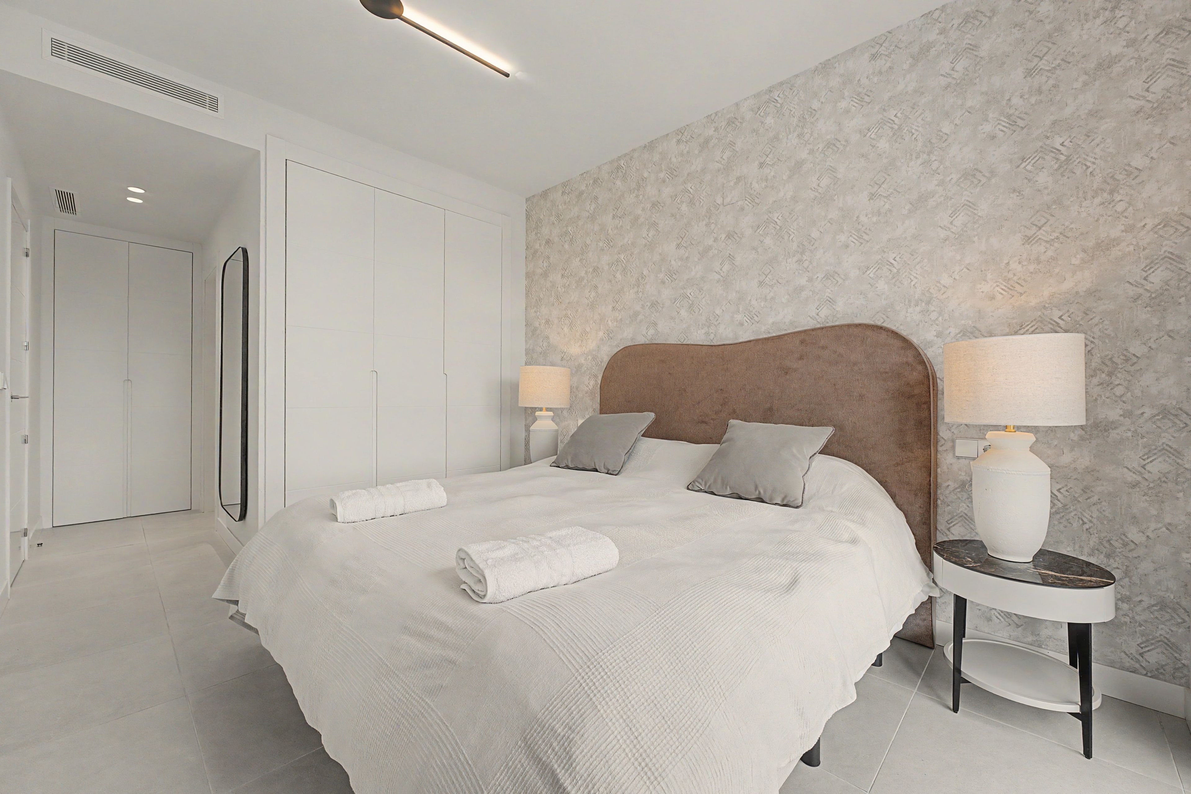 Un dormitorio moderno con una cama grande, dos mesitas de noche con lámparas, un armario y un papel tapiz estampado.