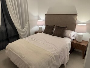 A neatly made bed with a headboard, two bedside lamps, and a window with curtains.