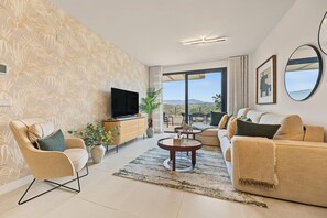 A modern living room with a flat-screen TV, a sofa, a coffee table, and a large window offering a view of the outdoors.