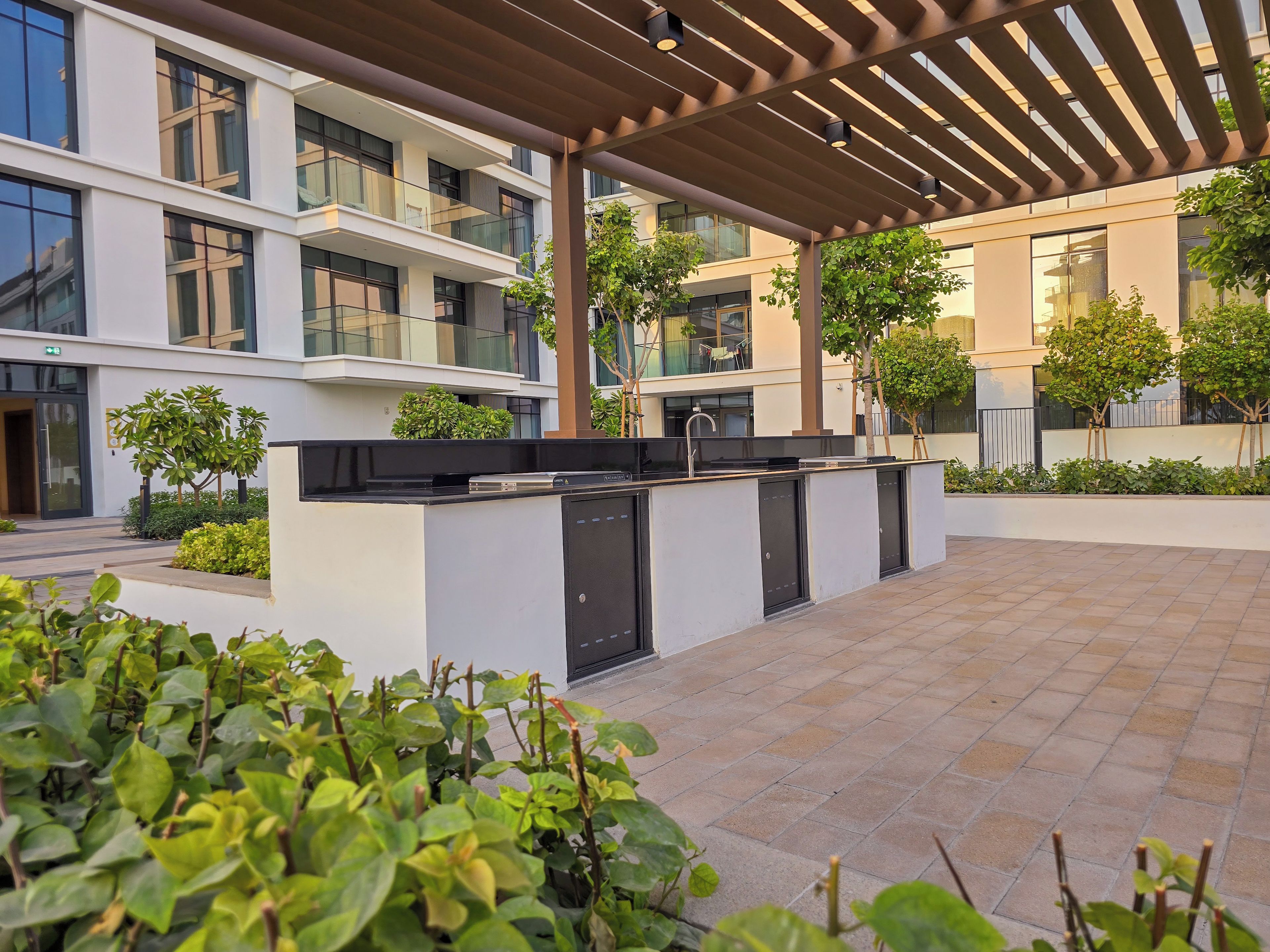 Un espace cuisine extérieur moderne, doté d’un barbecue encastré et d’un bar, entouré de verdure et agrémenté d’un patio couvert.