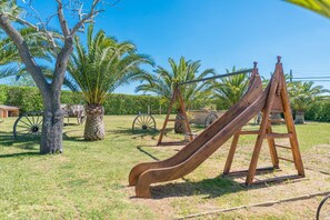 Un terrain de jeux avec un toboggan en bois et des balançoires, entouré de palmiers et surplombé d’un ciel dégagé.