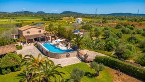 Ett stort hus med en swimmingpool, omgivet av frodig grönska och kullar i bakgrunden.